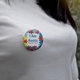 A close-up of a round badge pinned to a white shirt. The badge features a colourful jigsaw puzzle pattern with text reading 