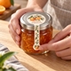 Hands applying an orange blossom lollipop seal sticker across the lid and front of a marmalade jar.
