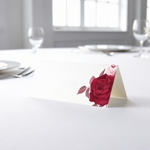 blank wedding place card with an elegant design printed on folded white cardstock, ready to write your names. Placed on a table set for a formal event, with plates, cutlery, and wine glasses in the softly blurred background.