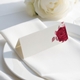 wedding place card with an elegant design on folded white cardstock, ready to write your names on. Positioned on a neatly folded white napkin atop a white plate, with a soft-focus white rose in the background.