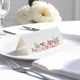 blank wedding place card with elegant design on folded white cardstock. Placed on a white plate with silver cutlery and a neatly folded napkin, with soft-focus white flowers in the background.