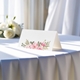 blank wedding place card printed with an elegantdesign on folded white cardstock ready to write your names on. Displayed on a table with a flowing white tablecloth, with soft natural light and white roses in the background.