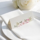 wedding place card with an elegant design on folded white cardstock, ready to write your names on. Positioned on a neatly folded white napkin atop a white plate, with a soft-focus white rose in the background.