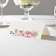 wedding place card with an elegant design on folded white cardstock ready to write your name on. Displayed on a white tablecloth with wine glasses, plates, and a floral centrepiece in the softly blurred background.