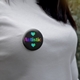 A close-up of a round badge pinned to a white shirt. The badge features a black background featuring the word 