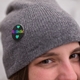 A close-up of a round badge pinned to a grey knit hat worn by a person. The badge displays a black background featuring the word 