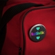 A round badge pinned to a red backpack. The badge features a a black background featuring the word 