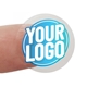 Transparent circle label sticker with a custom blue and white logo, shown adhered to a fingertip. The clear material allows the background to be visible, highlighting the personalised design.