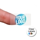 Transparent rectangle label sticker with a custom blue and white logo, shown adhered to a fingertip. The clear material allows the background to be visible, highlighting the personalised design.