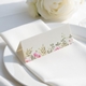 wedding place card with an elegant design on folded white cardstock, ready to write your names on. Positioned on a neatly folded white napkin atop a white plate, with a soft-focus white rose in the background.