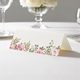 wedding place card with an elegant design on folded white cardstock ready to write your name on. Displayed on a white tablecloth with wine glasses, plates, and a floral centrepiece in the softly blurred background.