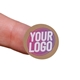 Kraft circle label sticker with a custom purple and white logo, shown on a fingertip. The natural brown background of the label gives a rustic and eco-friendly appearance, ideal for personalised packaging.