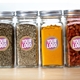 Four glass spice jars with silver lids, each labelled with a kraft circle sticker featuring a custom pink and white logo. The jars contain herbs, turmeric, and dried beans, showcasing personalised eco-friendly labels.