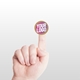 Small kraft circle label sticker with a custom pink and white logo, placed on the tip of an extended index finger. The minimalist white background accentuates the personalised design and eco-friendly material.