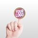 large kraft circle label sticker with a custom pink and white logo, placed on the tip of an extended index finger. The minimalist white background accentuates the personalised design and eco-friendly material