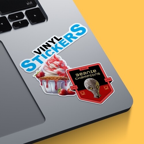 A collection of vinyl stickers placed on a grey laptop surface. The designs include a Vinyl Stickers label in bold blue text, a strawberry sundae with dripping sauce, and a red shield featuring an alien head with the text Beanie Champions and Top Secret.