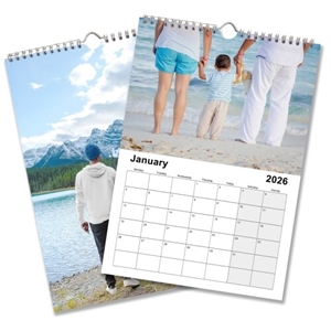 A 2026 wall calendar is shown open to January, featuring a family holding hands on a beach as the top image. The calendar grid below highlights the month of January with large, clear date boxes. Another page beneath shows a scenic image of a man standing by a mountain lake with snowy peaks and pine trees in the background. The calendar is wire-bound with a central hanging hook.