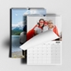 A 2025 A4 photo calendar featuring a family dressed as superheroes on the top page. The calendar is spiral-bound and partially open, revealing a blank month grid. Behind it, another image shows a person standing by a lake with mountains in the background.