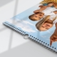 A close-up of a spiral-bound wall calendar lying flat on a white surface, showing a cover image of two smiling children—one older girl and one toddler—lying on straw against a bright blue sky with clouds. The metal hanging hook is clearly visible at the centre of the binding, and soft shadows suggest natural daylight coming through a nearby window.