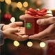 A close-up of a finger holding a round personalised Christmas sticker featuring elf legs in green shoes with red trim. The background is dark green with yellow stars, and the name Kit is written in white. A white gift with a red bow is partially visible.