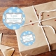 A personalised round Christmas label with a blue background and white snowflakes, featuring the message To James, Merry Christmas, Love Daddy. The label is shown on a brown paper-wrapped gift tied with white string, alongside an envelope.