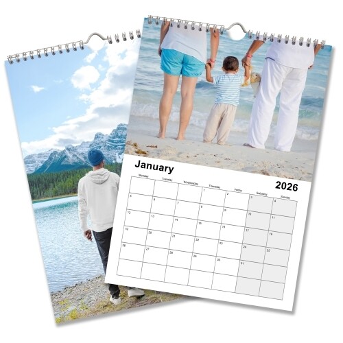 A 2026 wall calendar is shown open to January, featuring a family holding hands on a beach as the top image. The calendar grid below highlights the month of January with large, clear date boxes. Another page beneath shows a scenic image of a man standing by a mountain lake with snowy peaks and pine trees in the background. The calendar is wire-bound with a central hanging hook.