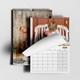 A 2026 wall calendar is shown with two pages visible. One page features a photo of a young child kissing a baby on a rustic white bench, partially turned to reveal a December calendar grid. Behind it, another page displays a festive scene with a smiling woman peeking out beside a wooden wall decorated with string lights and stars. The calendar is spiral-bound with a hanging hole at the top.