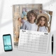 A 2026 wall calendar lies on a white surface open to April, featuring a photo of two children in white shirts, one wearing a straw hat, posing in front of pastel balloons. Behind it is another calendar page with a festive wooden background and a woman smiling next to fairy lights. A smartphone and pen rest nearby, suggesting a tidy desk or workspace setting. The calendar is spiral-bound with a hanging hook.