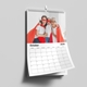 A 2026 wall calendar is displayed open to October, hanging on a white wall. The top image features three generations—an older woman, a younger woman, and a child—dressed as superheroes with red capes and masks, smiling and hugging. The calendar grid below shows the days of October 2026, with a simple, clean layout and large date boxes.