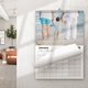 A 2026 wall calendar lies on a white surface open to April, featuring a photo of two children in white shirts, one wearing a straw hat, posing in front of pastel balloons. Behind it is another calendar page with a festive wooden background and a woman smiling next to fairy lights. A smartphone and pen rest nearby, suggesting a tidy desk or workspace setting. The calendar is spiral-bound with a hanging hook.