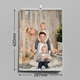 A wall calendar measuring 210mm wide by 297mm tall is shown hanging on a grey wall. The cover features a smiling family of four in cosy winter clothing, posing in front of a rustic wooden backdrop with warm fairy lights and festive decor. The calendar is wire-bound with a central hanging hook, ideal for A4 portrait display.