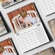 A 2026 wall calendar lies on a flat surface, open to July. The featured photo shows a young child lovingly cuddling a baby on a rustic white bench in front of a red door. The calendar grid below clearly displays the days of the month. Several other calendar pages with different photos and dates are scattered around, showing the product's variety and layout. The calendar is wire-bound with a central hanging hook.