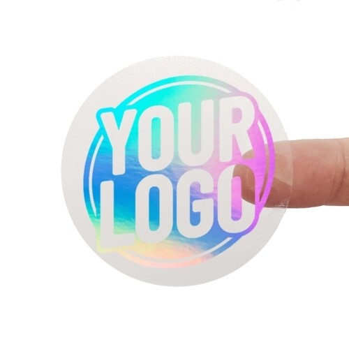 A transparent square sticker with rainbow holographic foil text reading Rainbow Holographic Foiled Stickers. A person's finger is visible behind the sticker, showing its clear background. The foil lettering reflects different colours depending on the angle.