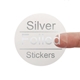 A transparent square sticker with silver foil text reading Silver Foiled Stickers. A person's finger is visible behind the sticker, showing its clear background. The silver foil lettering has a reflective metallic finish for a premium look.