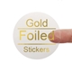 A transparent square sticker with gold foil text reading Gold Foiled Stickers. A person's finger is visible behind the sticker, demonstrating its clear background. The gold foil lettering adds a premium and reflective finish.