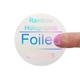 A transparent square sticker with rainbow holographic foil text reading Rainbow Holographic Foiled Stickers. A person's finger is visible behind the sticker, showing its clear background. The foil lettering reflects different colours depending on the angle.