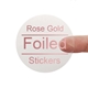 A transparent square sticker with rose gold foil text reading Rose Gold Foiled Stickers. A person's finger is visible behind the sticker, showing its clear background. The foil lettering has a metallic sheen, creating an elegant and premium look.