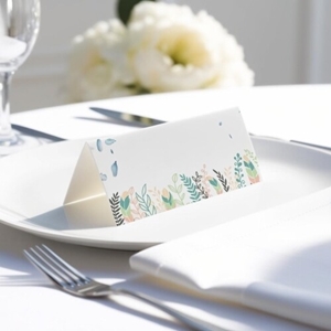 blank wedding place card with elegant design on folded white cardstock. Placed on a white plate with silver cutlery and a neatly folded napkin, with soft-focus white flowers in the background.