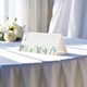 blank wedding place card printed with an elegantdesign on folded white cardstock ready to write your names on. Displayed on a table with a flowing white tablecloth, with soft natural light and white roses in the background.