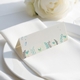 wedding place card with an elegant design on folded white cardstock, ready to write your names on. Positioned on a neatly folded white napkin atop a white plate, with a soft-focus white rose in the background.