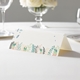 wedding place card with an elegant design on folded white cardstock ready to write your name on. Displayed on a white tablecloth with wine glasses, plates, and a floral centrepiece in the softly blurred background.