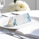 blank wedding place card with elegant design on folded white cardstock. Placed on a white plate with silver cutlery and a neatly folded napkin, with soft-focus white flowers in the background.