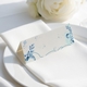 wedding place card with an elegant design on folded white cardstock, ready to write your names on. Positioned on a neatly folded white napkin atop a white plate, with a soft-focus white rose in the background.