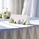 blank wedding place card printed with an elegantdesign on folded white cardstock ready to write your names on. Displayed on a table with a flowing white tablecloth, with soft natural light and white roses in the background.