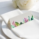 wedding place card with an elegant design on folded white cardstock, ready to write your names on. Positioned on a neatly folded white napkin atop a white plate, with a soft-focus white rose in the background.