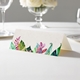 wedding place card with an elegant design on folded white cardstock ready to write your name on. Displayed on a white tablecloth with wine glasses, plates, and a floral centrepiece in the softly blurred background.