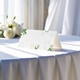 blank wedding place card printed with an elegantdesign on folded white cardstock ready to write your names on. Displayed on a table with a flowing white tablecloth, with soft natural light and white roses in the background.