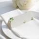 wedding place card with an elegant design on folded white cardstock, ready to write your names on. Positioned on a neatly folded white napkin atop a white plate, with a soft-focus white rose in the background.