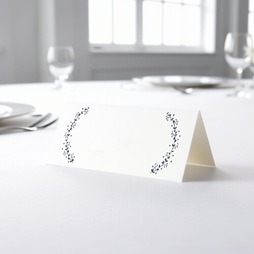 blank wedding place card with an elegant design printed on folded white cardstock, ready to write your names. Placed on a table set for a formal event, with plates, cutlery, and wine glasses in the softly blurred background.