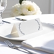 blank wedding place card with elegant design on folded white cardstock. Placed on a white plate with silver cutlery and a neatly folded napkin, with soft-focus white flowers in the background.